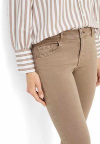 Jeans stretch beige con design aderente, caratterizzati da una piccola tasca frontale e una leggera texture del tessuto. Abbinati a una blusa a righe.