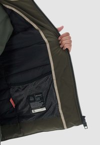 Interno della giacca verde oliva, con isolamento nero e una tasca in rete con un'etichetta di cura. Apertura con zip bordata in beige a contrasto.