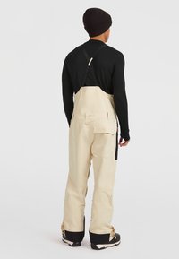 Pantalones de tirantes impermeables beige con tirantes negros, corte holgado, bolsillos laterales y dobladillo inferior reforzado. Llevados sobre una camiseta de manga larga negra.