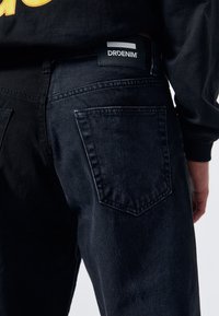 Černé denimové džíny s detailním pohledem, zahrnující zadní kapsu, černou a bílou značkovou visačku a jemně texturovanou látku.