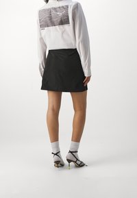 Chemise blanche boutonnée avec un grand motif gris au dos, associée à une jupe noire et des talons noirs à lanières avec des accents verts.