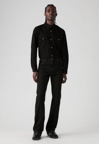 Zwart denim overhemd met twee borstzakken, gecombineerd met donker groene spijkerbroek. De outfit heeft gouden knopen en een rechte, aangesloten pasvorm.