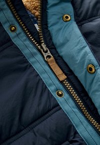 Close-up van een marineblauwe en lichtblauwe jasrits die gedeeltelijk open is, waardoor de metalen ritskammen, de stoffen trekker en de drukknoopjes zichtbaar zijn.