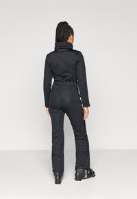 Μαύρη στολή σκι με ψηλό γιακά, υφασμάτινη υφή, φερμουάρ στα πόδια και λογότυπο στη ζώνη. Φοράει μαύρες μπότες σκι.