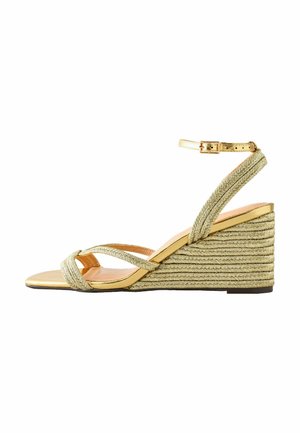 Kilklackssandal med flätad guldrem och spänne vid ankeln. Har en vävd juteklack, platt framdel och slät beige innersula.