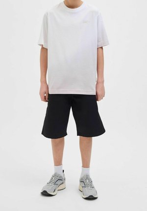 Personne portant un t-shirt blanc uni oversize, un short noir jusqu'aux genoux, des chaussettes blanches et des baskets grises, debout sur un fond blanc.