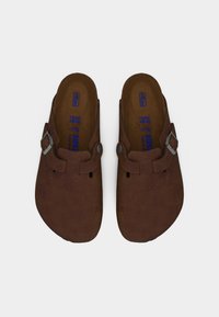 Καφέ μοκασίνια Birkenstock από σουέτ με ρυθμιζόμενες μεταλλικές αγκράφες και διαμορφωμένα πέλματα, απεικονίζονται από πάνω σε λευκό φόντο.