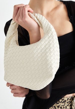 Sac à main blanc tissé avec une forme arrondie, une surface texturée et une poignée courte, tenu par une personne portant un haut noir. Fermeture zippée visible.