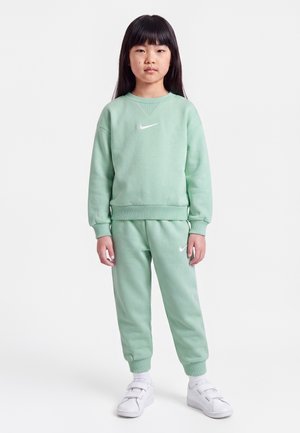 Jeune fille debout vêtue d'un sweat-shirt Nike vert menthe et d'un pantalon de survêtement avec des baskets blanches, sur un fond blanc uni.