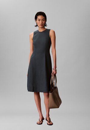 Donna con abito senza maniche grigio scuro fino al ginocchio, che tiene una borsa taupe, indossa sandali neri con tacco, in piedi davanti a uno sfondo grigio.