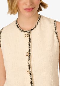 Krémový top bez rukávů s texturovanou tkaninou, s černým lemem a třemi zlatými knoflíky s perlovými akcenty podél předního zapínání.