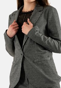 Blazer en blanco y negro con patrón de espiga y un ajuste entallado, con solapas de muesca y dos bolsillos frontales, llevado sobre una blusa de encaje negro transparente.