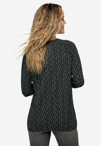 Zwart langek mouwtop met een herhalend groen tekstpatroon, ronde halslijn en een losse pasvorm, gecombineerd met grijze broek.