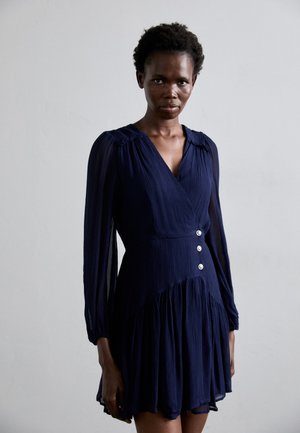 Robe portefeuille bleu marine avec de longues manches transparentes, des épaules froncées et trois boutons en argent à la taille ; elle présente une jupe évasée.