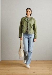 Giacca bomber verde con dettagli a zip e bottoni, jeans blu chiaro e sneakers bianche, mentre porta una borsa a tracolla crema. Tessuto morbido, vestibilità casual.