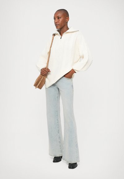 Suéter blanco de punto trenzado con cuello de cremallera, combinado con jeans acampanados de color azul claro y botas negras hasta el tobillo, junto a un bolso cruzado marrón.
