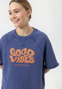 Blauwe sweatshirt met korte, opgerolde mouwen. Bevat oranje tekst: "GOOD VIBES" en "STATE OF MIND." Zacht, textuurstof met een ronde hals.