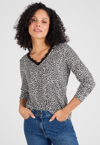 Haut à manches longues imprimé léopard avec un col en V et des bordures en dentelle. Fabriqué en tissu doux dans un schéma de couleurs crème et noir. Porté avec un jean bleu.