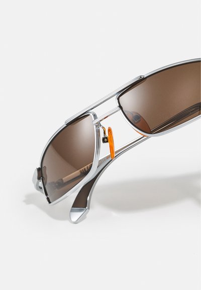 Lunettes de soleil argentées avec des verres marron, une monture métallique épurée et un détail orange. Présente un design de pont unique et une structure minimaliste.