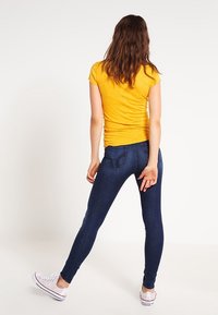 Žlutý krátký rukáv, upnutý vršek, tmavě modré skinny džíny se zadními kapsami, nošeno s bílými plátěnými teniskami, pohled zezadu.
