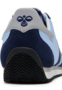 Sapatilha desportiva com parte superior em camurça e malha azul, capinha traseira cinza, sola de borracha preta com padrão em zig-zag e logótipo bordado na parte de trás.