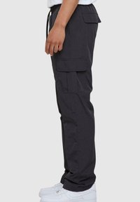 Nákladní kalhoty tmavě šedé látky, s bočními kapsami a elastickým pasem. Kombinované s bílými teniskami, zobrazeno ze strany.