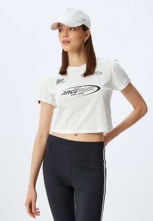Witte cropped t-shirt met grafische prints en tekst, gecombineerd met zwarte leggings met witte zijstrepen. Lichte stof, korte mouwen.