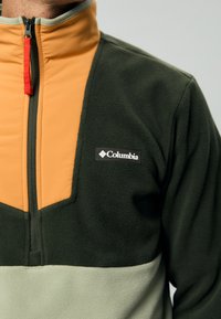 Muškarac nosi Columbia flis jaknu sa zelenim, narančastim i svijetlo sivim blokovima boja, s polu-zip zatvaranjem i crvenim povlačenjem.