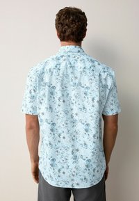 Camisetta a maniche corte di colore azzurro chiaro con motivo floreale. Presenta un colletto classico, chiusura con bottoni e orlo arrotondato.