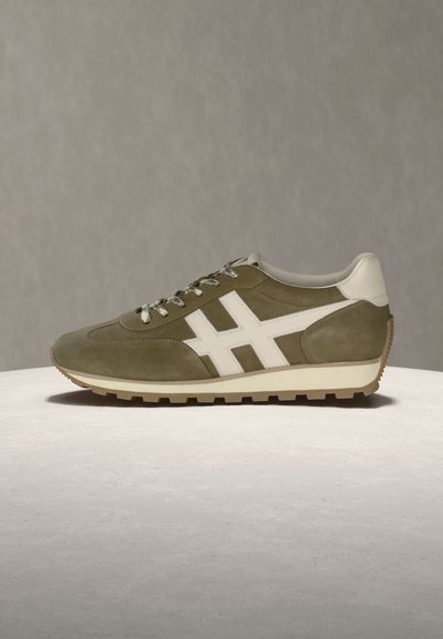 Baskets en daim vert olive avec accents en cuir blanc, lacets à motifs et semelle en caoutchouc beige robuste sur une surface et un fond neutres.