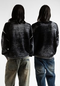 Δύο άτομα που φορούν υπερμεγέθη, μαύρα και γκρι gradient πουλόβερ με υφασμάτινη υφή και ραμμένες μανσέτες, συνδυασμένα με ανοιχτόχρωμα και σκούρα τζιν.