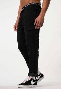 Schwarze Cargo-Hosen mit einem Kordelzugbund und Seitentaschen, aus strukturiertem Stoff. Kombiniert mit schwarz-weißen Nike-Sneakern.
