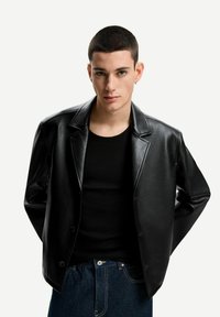 Schwarze Kunstlederjacke mit Reverskragen, Knopfverschluss und lockerer Passform. Über einem schwarzen Hemd getragen, kombiniert mit dunklen Jeans.