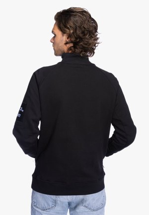 Man with wavy hair wearing a black high-neck sweatshirt and light blue jeans, standing with hands on hips, facing away.