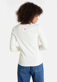 Frau mit lockigem Haar, die ein langärmliges weißes Hemd und eine blaue Jeans trägt, von hinten vor einem weißen Hintergrund gezeigt.