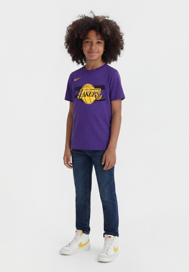 Child with curly hair wearing a purple Los Angeles Lakers T-shirt, blue jeans, and white sneakers with yellow Nike swoosh on a white background.