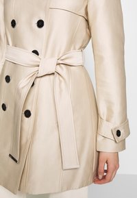 Beige trench coat s páskem s hedvábnou texturou, který má černé knoflíky, dvouřadové zapínání a upravený střih na manžetách.