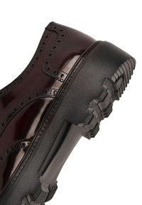 Zapato de charol burdeos con detalles perforados y una gruesa suela de goma negra. Presenta estrías distintivas y un perfil elegante.