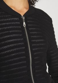 Close-up of a person wearing a black textured knit jacket with horizontal patterns and a central silver zipper, collar visible.