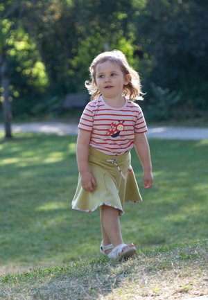 Child wearing a red and white striped t-shirt with a snail graphic, paired with a green skirt. Standing on grass, with sunlight filtering through.