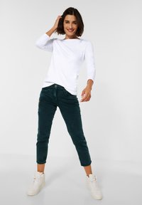 Witte lange mouwen shirt gecombineerd met donker groene corduroy broek tot de enkels en witte hoge sneakers, die een casual outfit tonen.