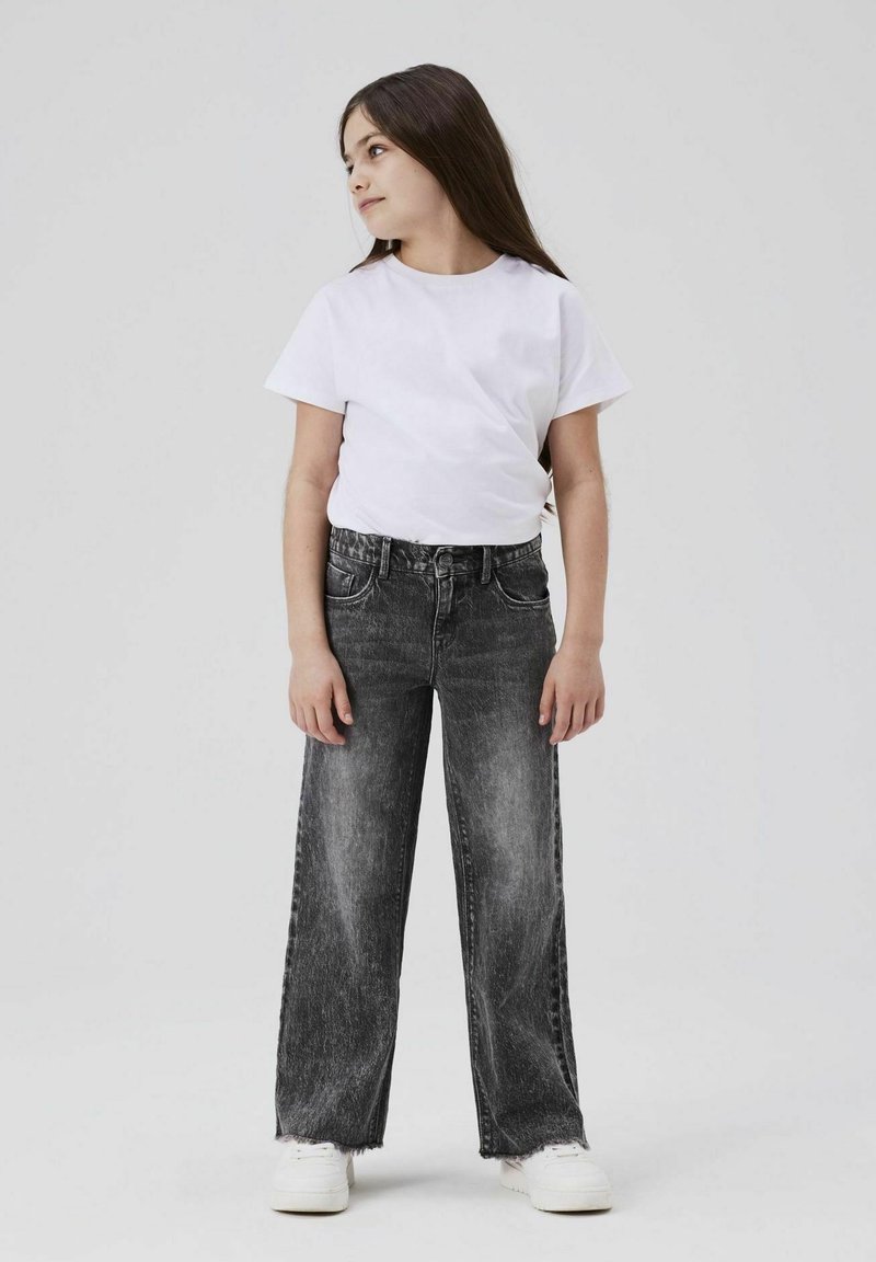Una camiseta blanca de manga corta, combinada con unos jeans de pierna ancha de talle alto en negro con un acabado desgastado y dobladillos deshilachados, usados con zapatillas blancas.
