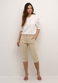 Top mit Puffärmeln in Creme, kombiniert mit knielangen beigen Hosen, die über Vorderaschen und eine schmale Passform verfügen. Das Outfit wird durch braune Sandalen mit Riemchen ergänzt.