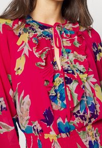 Jasně růžová blůza s květinovými vzory, volány podél výstřihu a uzlem v klíčové díře. Vyrobena z lehké, texturované tkaniny.