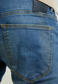 Jean en denim bleu avec une poche arrière à la forme angulaire unique et des coutures jaunes, complété par une étiquette de marque en cuir foncé.