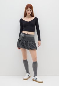 Schwarzes Langarmshirt, grauer Rock mit Rüschen und einem Gürtel, graue Kniestrümpfe und weiße Schuhe mit marineblauen Akzenten.