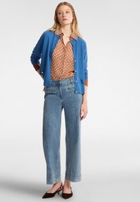 Cardigan blu su una camicia a fantasia marrone, abbinato a jeans a gamba larga celesti e scarpe marroni. Caratterizzato da bottoni e polsini a contrasto.