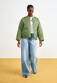 Podložena olivno zelena jakna s okroglim ovratnikom, pritisnimi gumbi in sprednjim žepom, v kombinaciji z širokima svetlim denim hlačami in mornarsko modrimi teniskami.