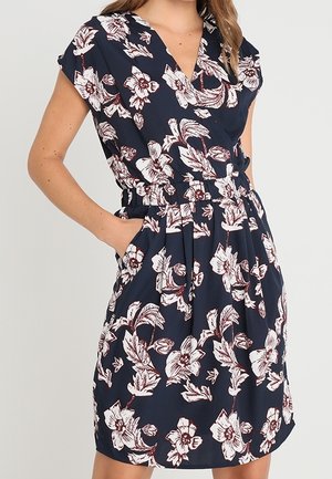 Femme portant une robe portefeuille bleu marine avec des motifs floraux blancs et bordeaux, manches courtes et poches aux hanches.