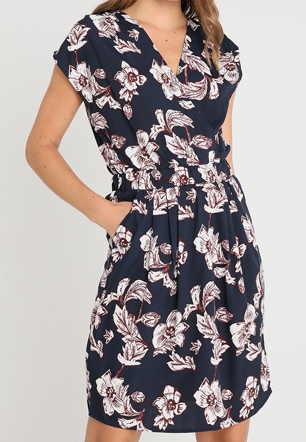 Femme portant une robe portefeuille bleu marine avec des motifs floraux blancs et bordeaux, manches courtes et poches aux hanches.